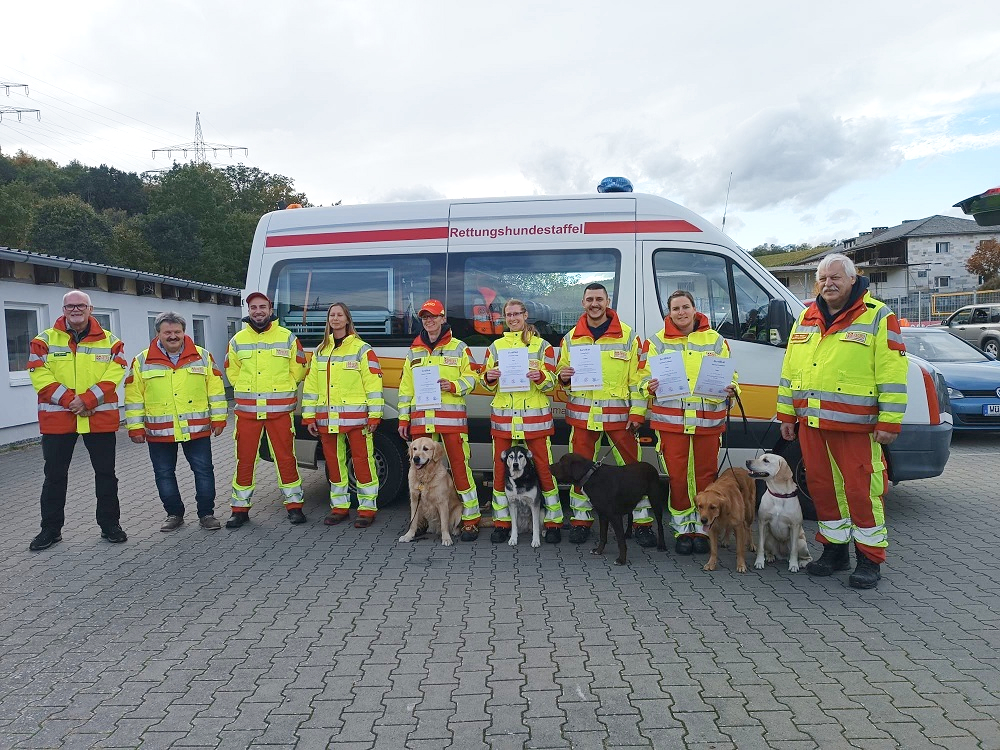 9 Rettungshundeführer stehen mit 4 Hunden nebeneinander in gelb-roter Schutzkleidung vor dem Einsatzfahrzeug
